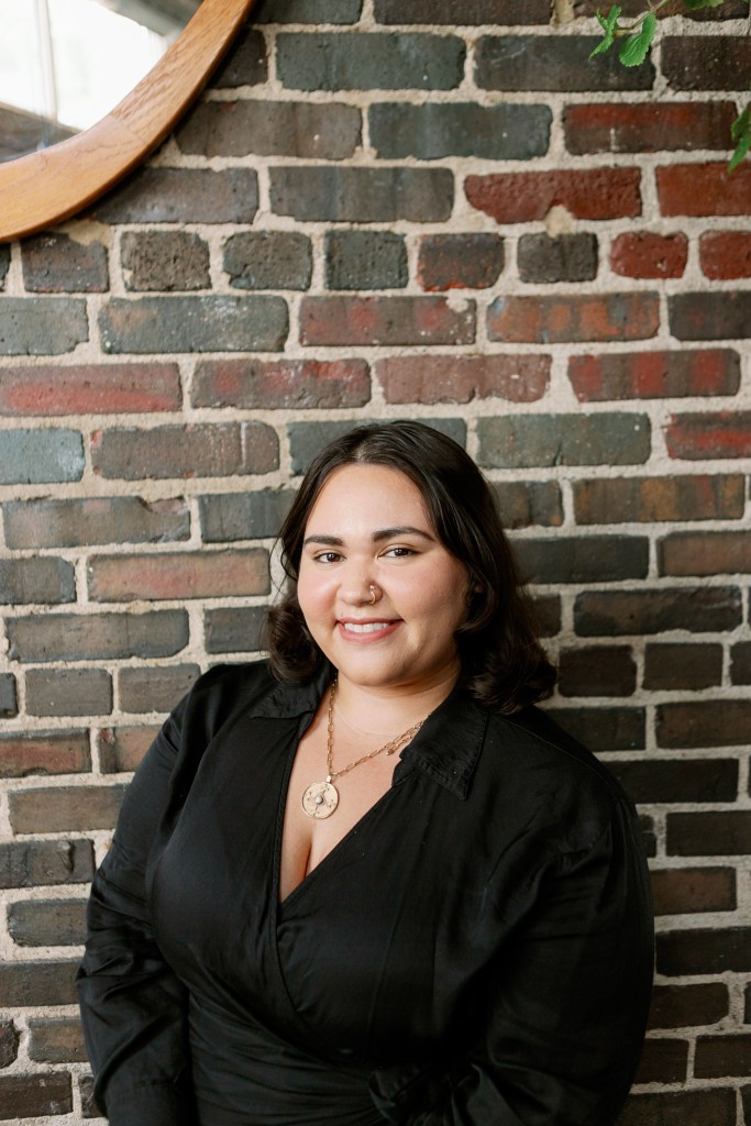 A woman with dark, wavy hair smiles while wearing a black outfit, standing against a rustic brick wall with a round wooden mirror in the background.